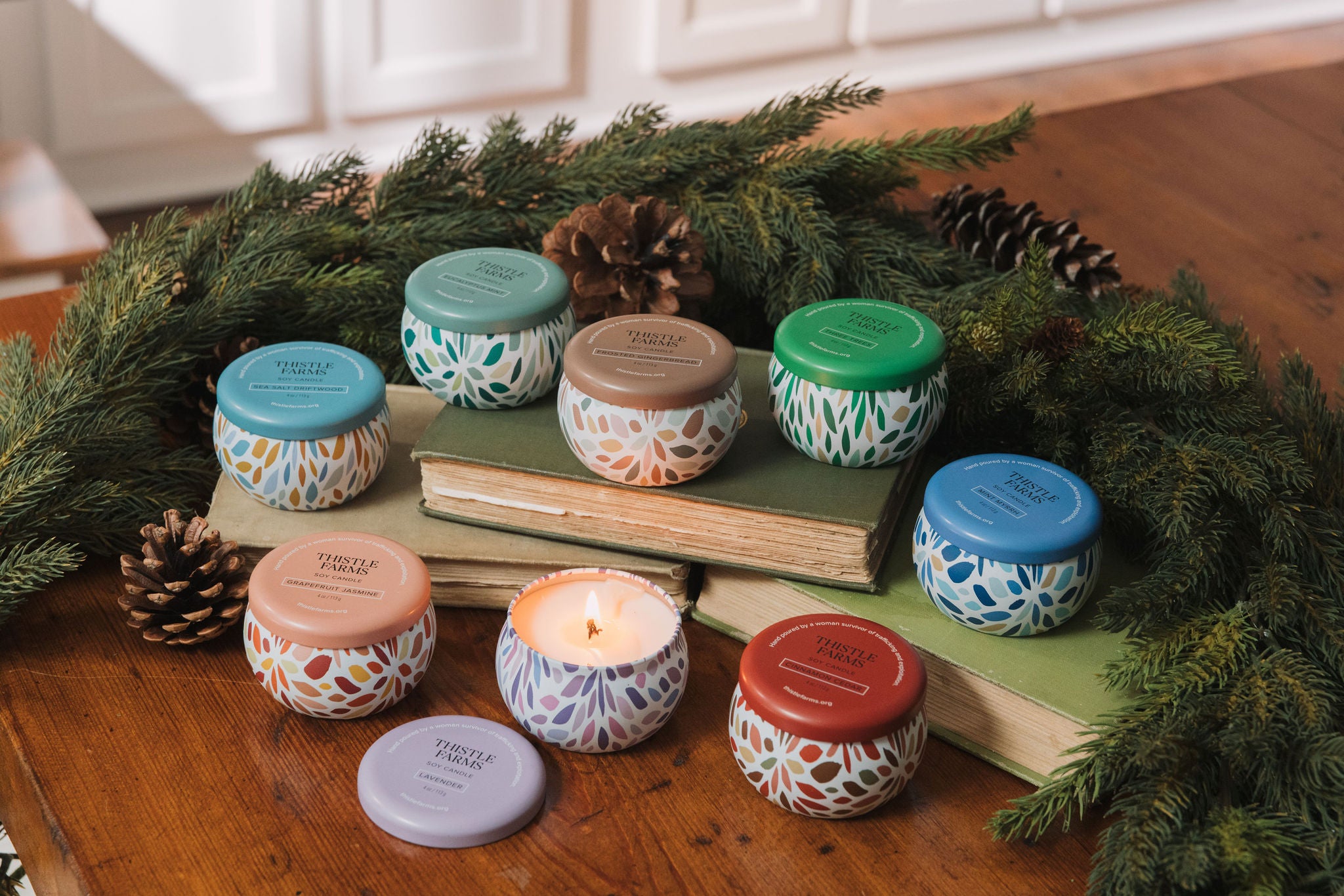 Assorted candles with colorful lids on a wooden surface with greenery and books.