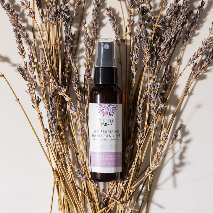Bottle of Thistle Farms hand sanitizer surrounded by lavender flowers on a light background