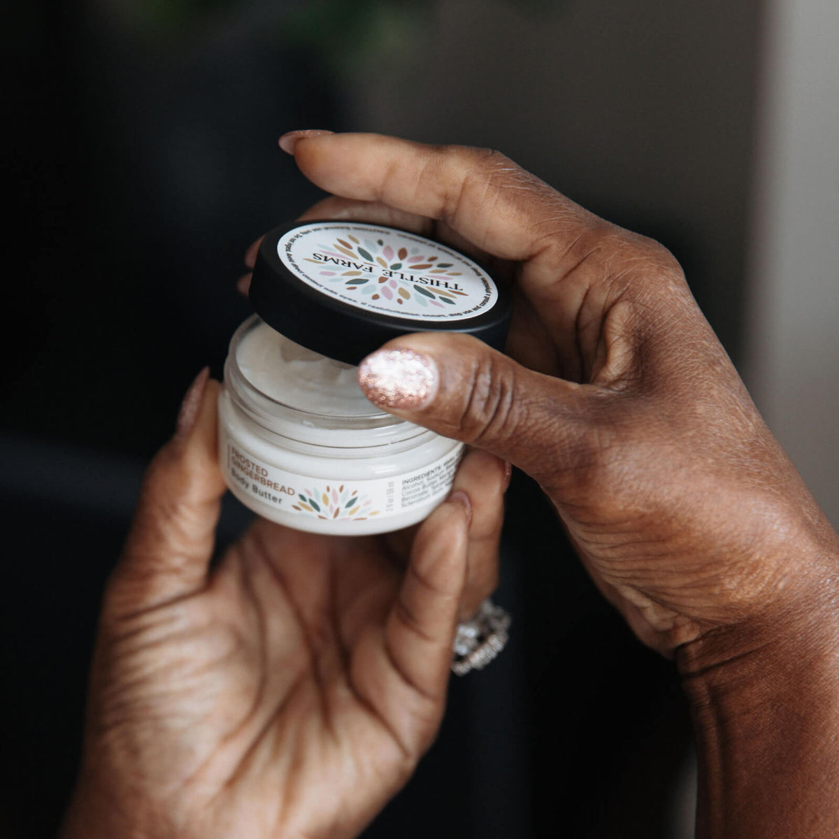 Hand holding a small jar of body butter against a dark background