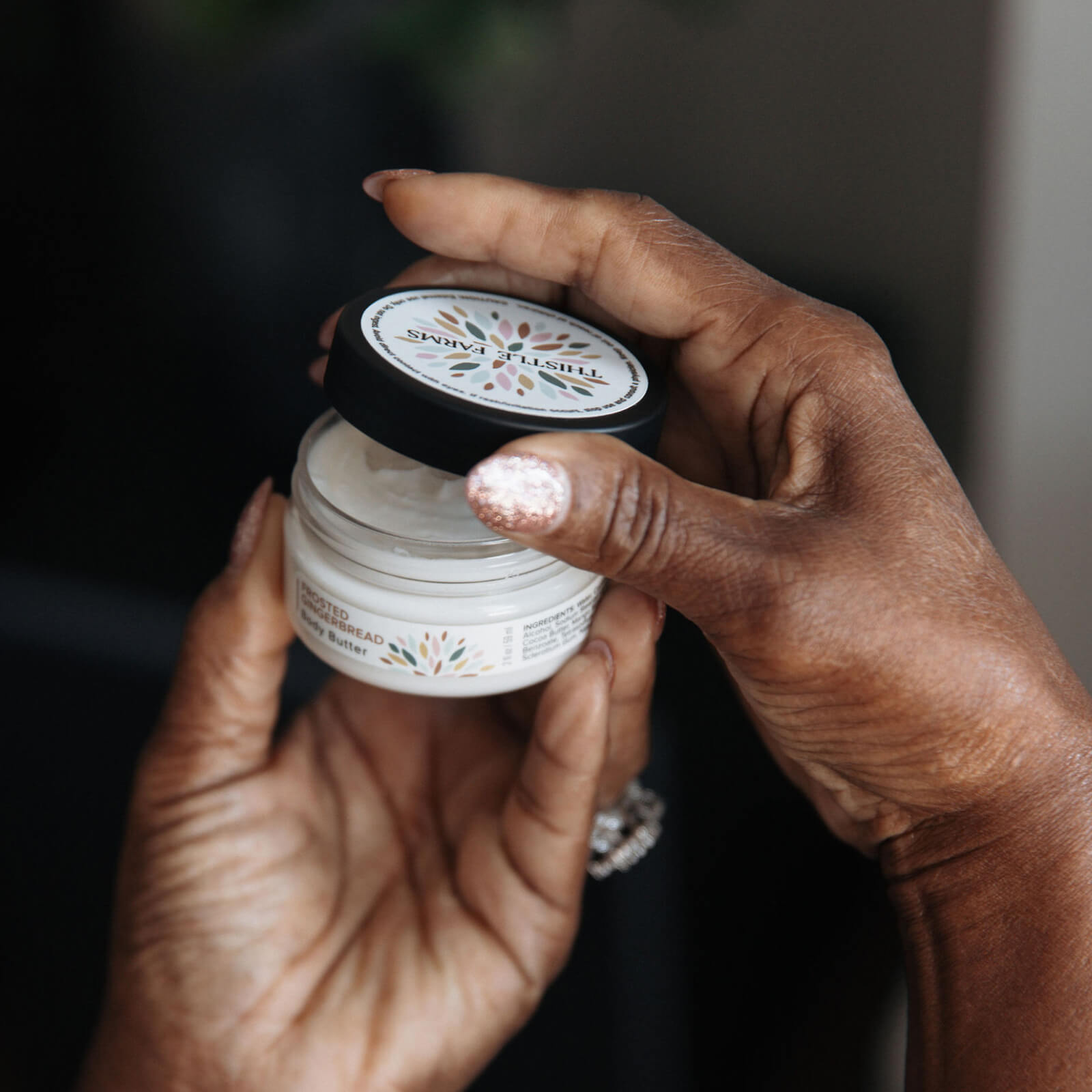 Hand holding a small jar of body butter against a dark background