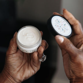 Two hands holding small jars of cream against a dark background