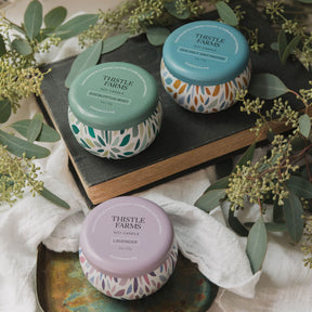Three Thistle Farms candles on a wooden surface with greenery around