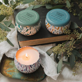 Three candles with decorative lids on a textured surface with greenery
