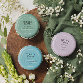Three Thistle Farms candles on a wooden board with greenery and flowers.