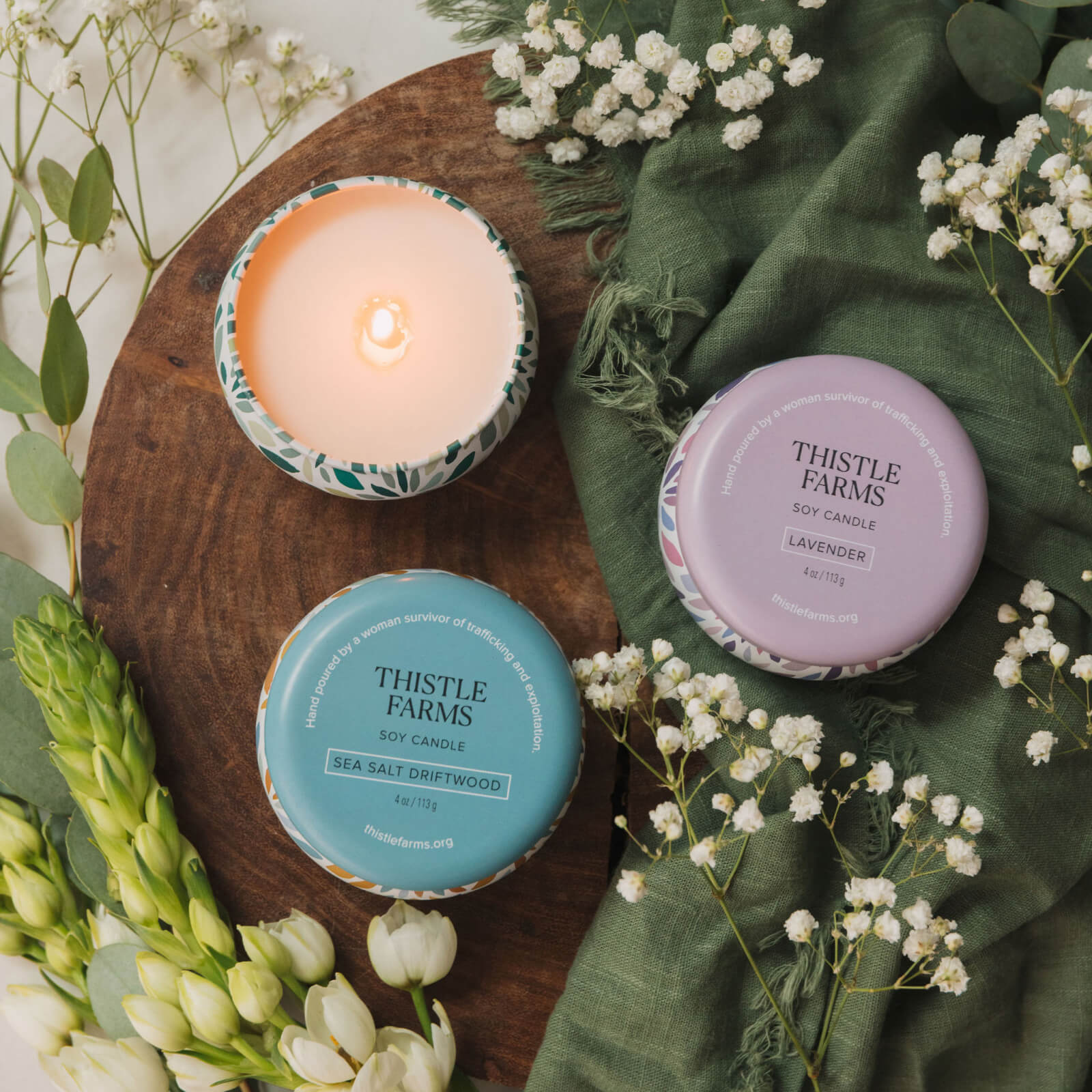 Three Thistle Farms candles on a wooden board with flowers around