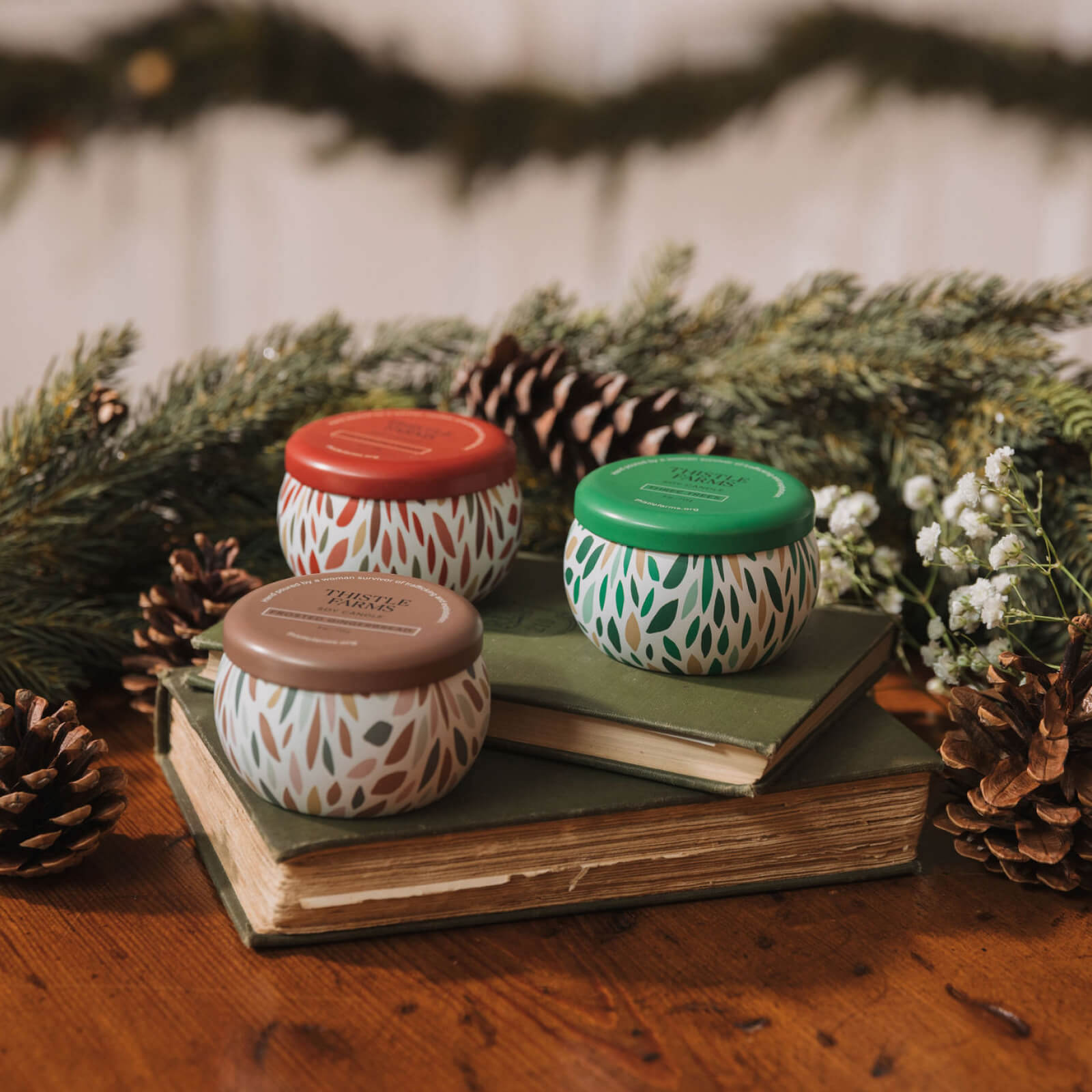 Three small decorative jars with patterned lids on a book surrounded by pine cones and greenery.