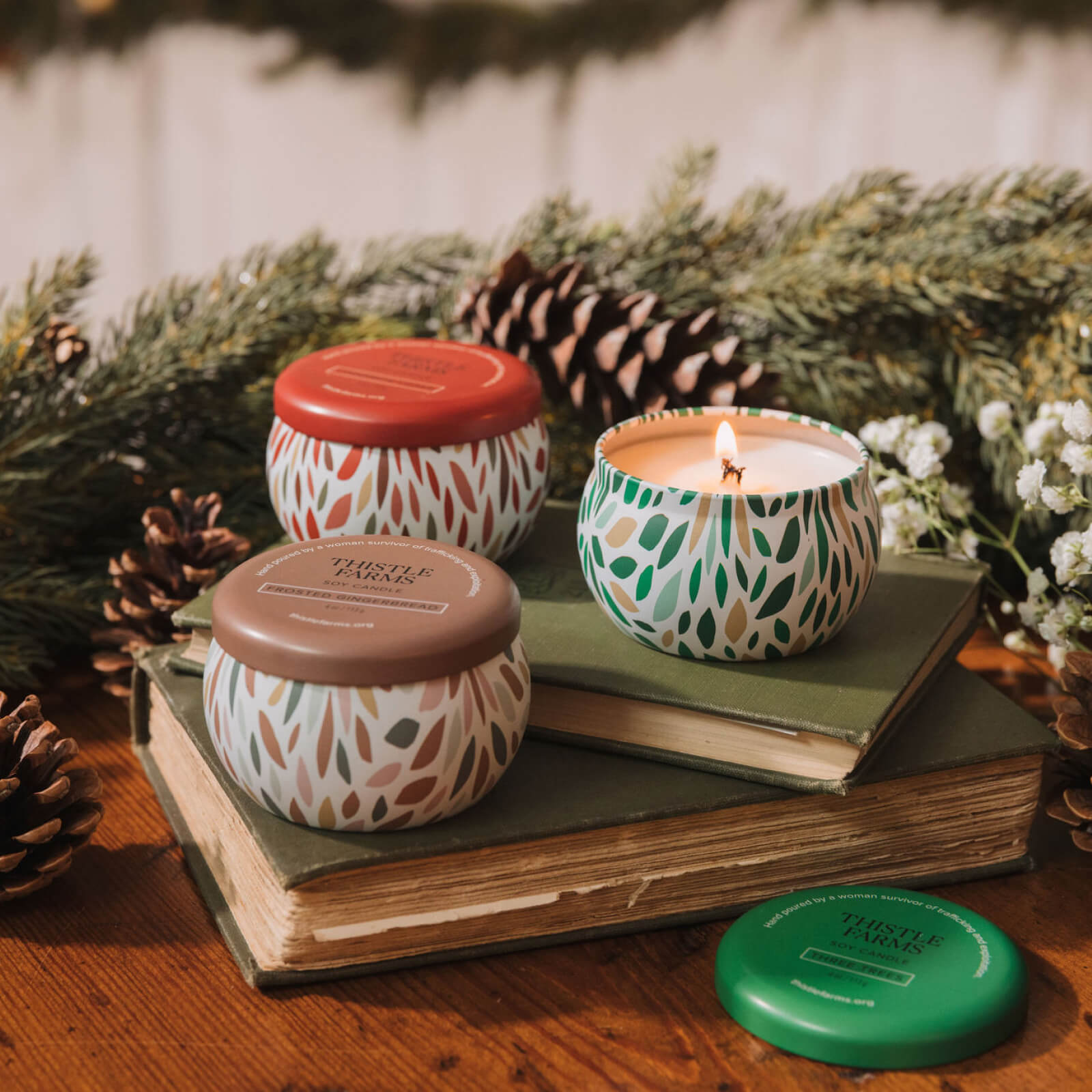 Decorative candles with patterned lids on books, surrounded by greenery and pinecones.