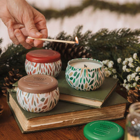Hand lighting a candle with decorative lids on a stack of books, surrounded by greenery.