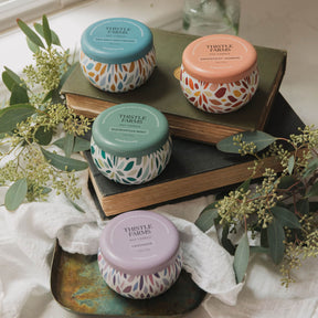 Five small jars with colorful lids on books, surrounded by greenery and fabric.