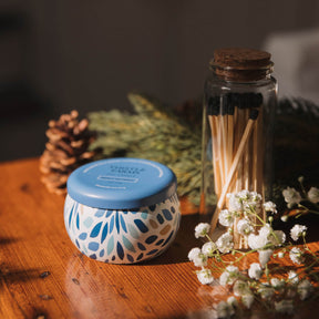 Candle with blue lid, matches in a jar, and small plants on a wooden surface