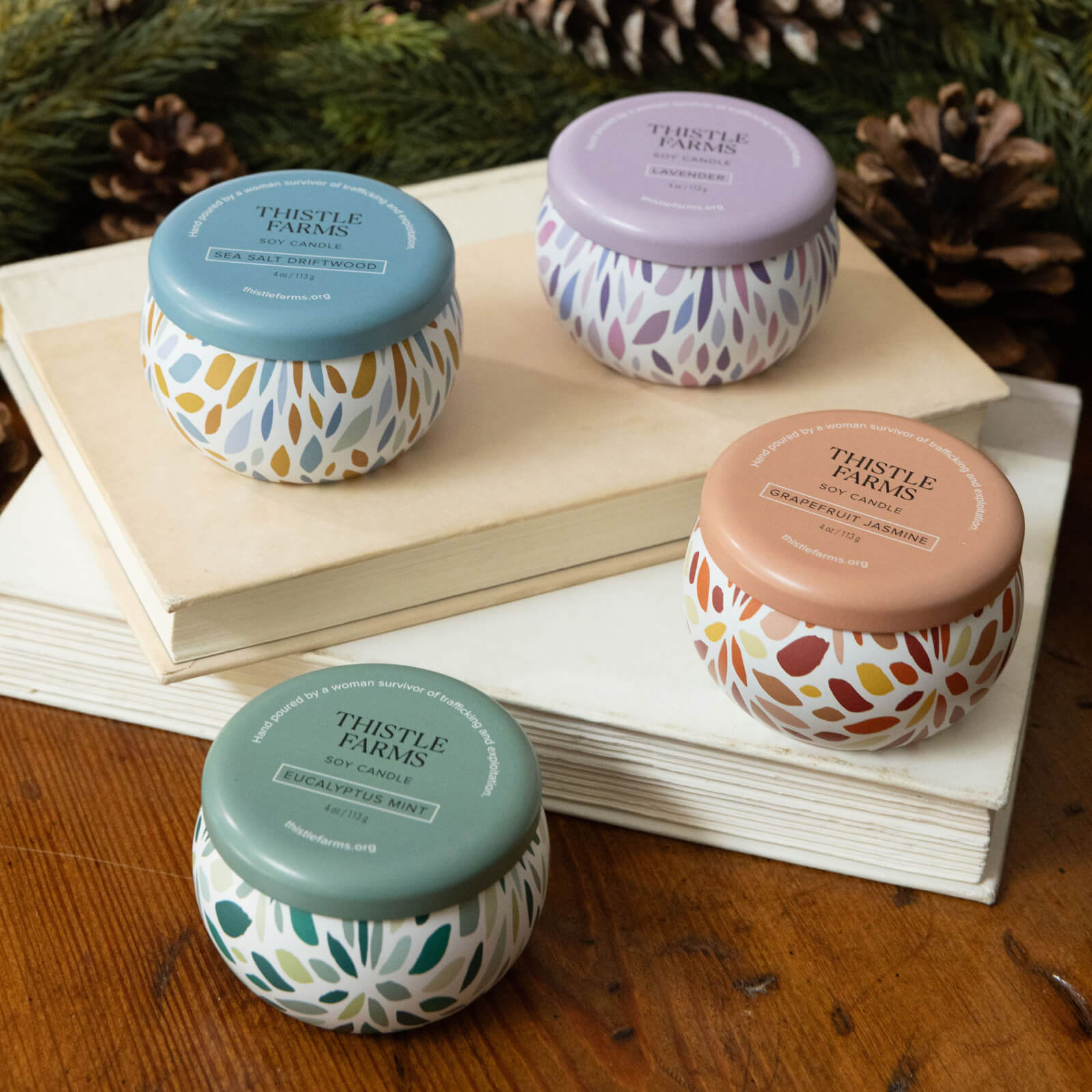 Four Thistle Farms candles with colorful lids on a wooden surface with books and pine cones in the background.