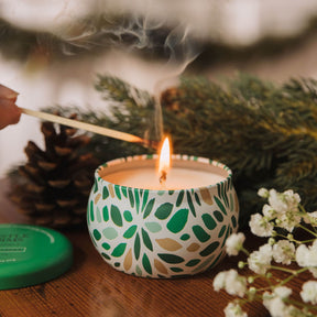Candle with a pattern being lit next to pine cones and flowers on a wooden surface