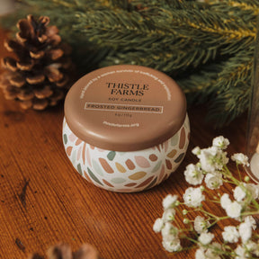 Thistle Farms soy candle with brown lid on a wooden surface with greenery and flowers.