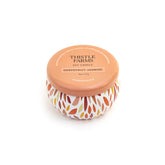 Thistle Farms soy candle with an orange lid and a patterned base on a white background