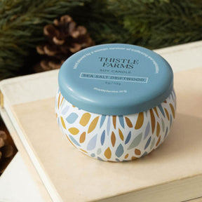 Thistle Farms soy candle with a blue lid on a wooden surface with greenery in the background